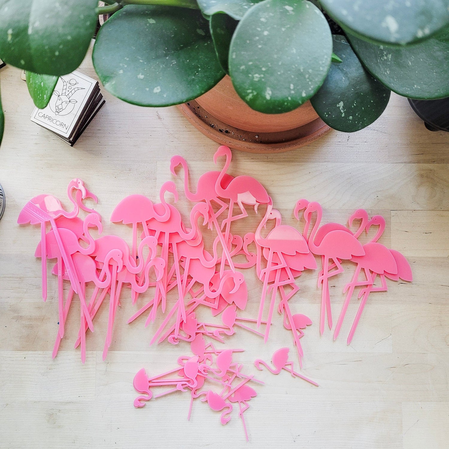 Pile of mini pink flamingo plant stakes on a light wood desktop with a plant in the background.