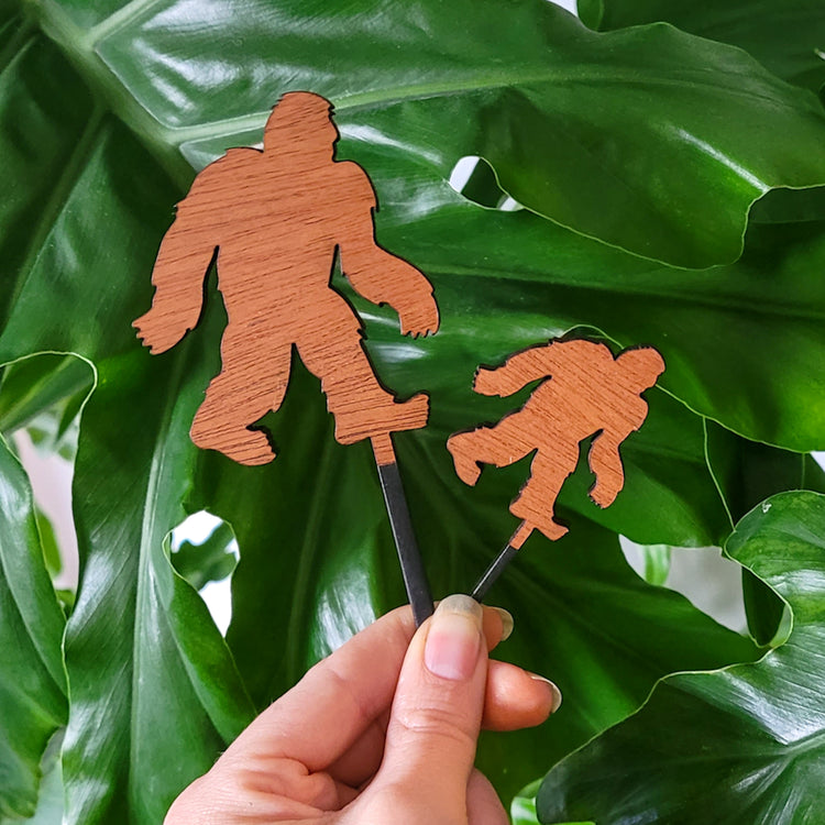 Bigfoot silhouette decorative plant stakes in two sizes held in hand with philodendron leaves in the background.