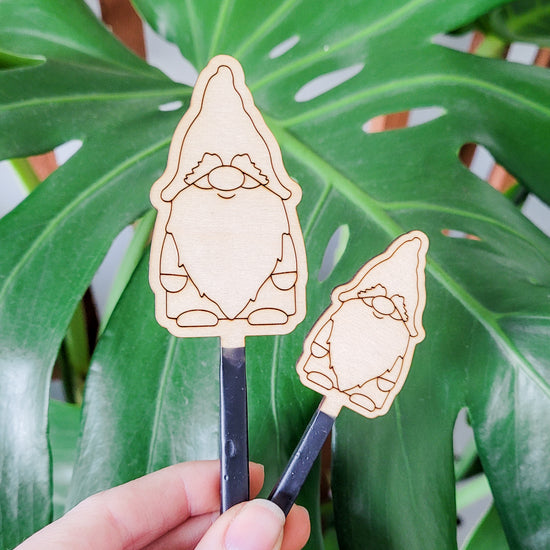 Decorative plant stake accessories featuring a garden gnome engraved in light wood. Both size options held in hand with monstera leaf in the background.