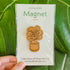 Handcrafted magnet featuring a fiddle leaf fig plant engraved in cherry wood held against a plant in product packaging.