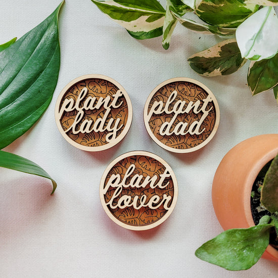 Three handcrafted wooden magnets with the text "Plant Lady," "Plant Lover," and "Plant Dad" cut from light colored wood on a darker wood background with an engraved monstera leaf pattern. 