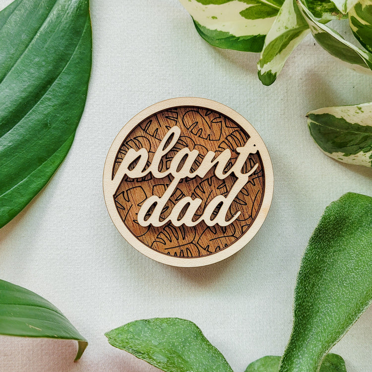Handcrafted wooden magnet with the text "Plant Dad" cut from light colored wood on a darker wood background with an engraved monstera leaf pattern. 