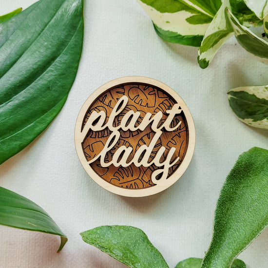 Handcrafted wooden magnet with the text "Plant Lady" cut from light colored wood on a darker wood background with an engraved monstera leaf pattern. 