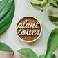 Handcrafted wooden magnet with the text "Plant Lover" cut from light colored wood on a darker wood background with an engraved monstera leaf pattern. 