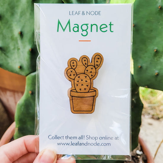 Handcrafted magnet featuring an opuntia cactus engraved in cherry wood held against a plant in product packaging.