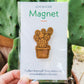 Handcrafted magnet featuring an opuntia cactus engraved in cherry wood held against a plant in product packaging.